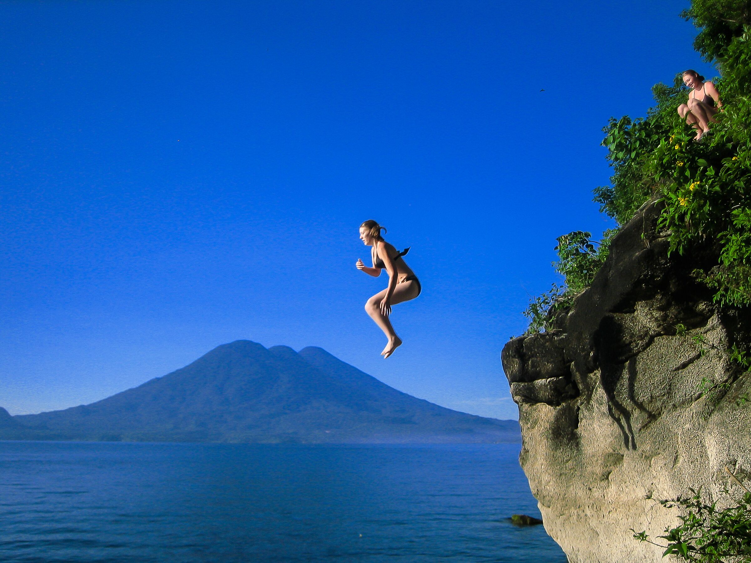 Una persona salta da una scogliera in acqua, con una montagna sullo sfondo.