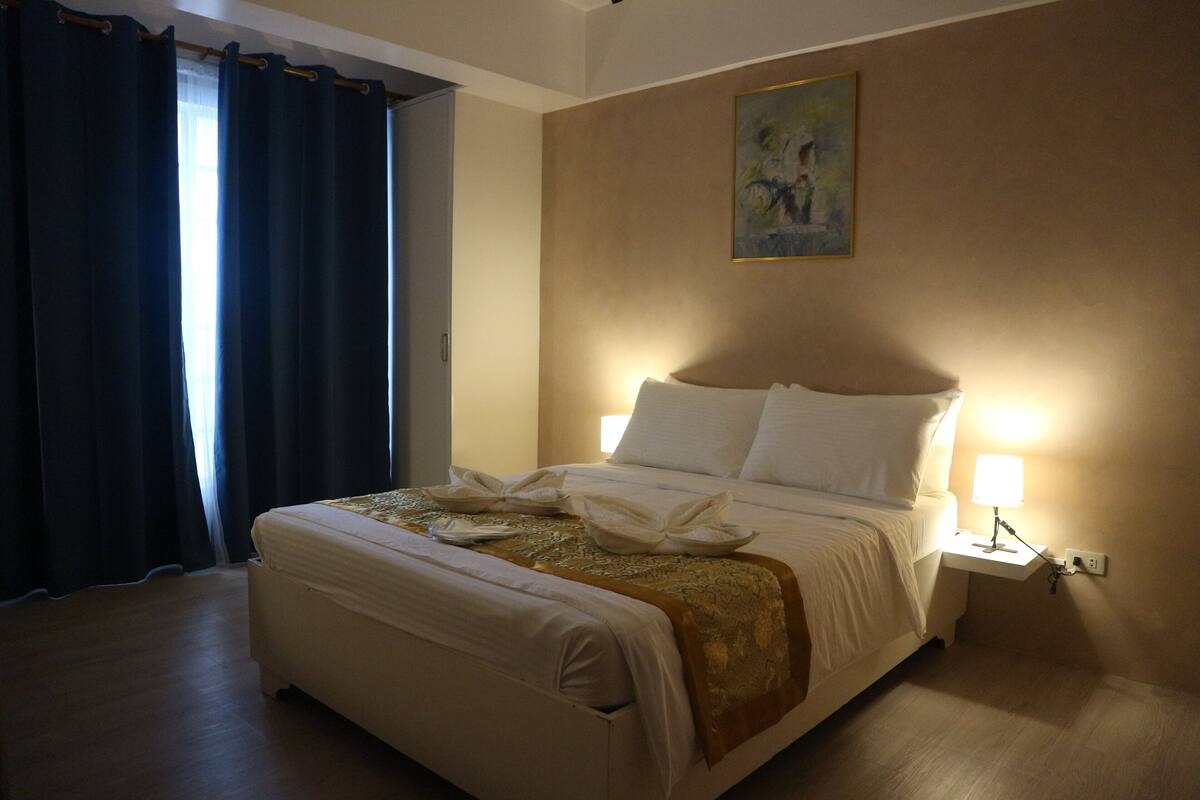 Una cama bien hecha con sábanas blancas y un edredón dorado, una mesita de noche con lámpara, y un cuadro en la pared.