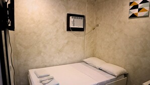 A small, single-bed room with a white bed, a wall-mounted air conditioning unit, and a plain wall with a geometric-patterned artwork. - Hotel Casa Ilustre (Balayan)