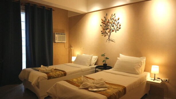 A hotel room with two beds, a wall-mounted air conditioner, a small table with a lamp, and a plant on the wall. - Hotel Casa Ilustre (Balayan)