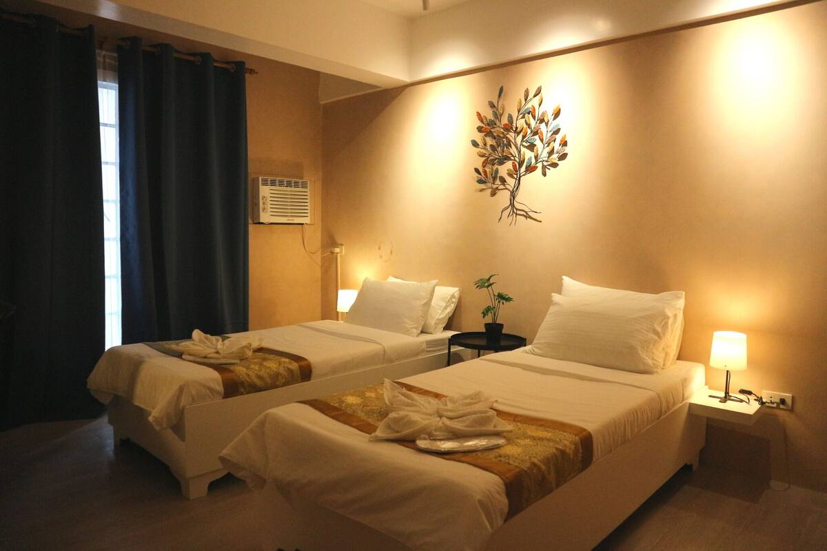 Habitación de hotel con dos camas, aire acondicionado instalado en la pared, una mesita con lámpara y una planta en la pared.