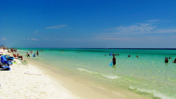 Una playa con agua turquesa cristalina, gente nadando y tomando el sol, y sillas y sombrillas en la arena.