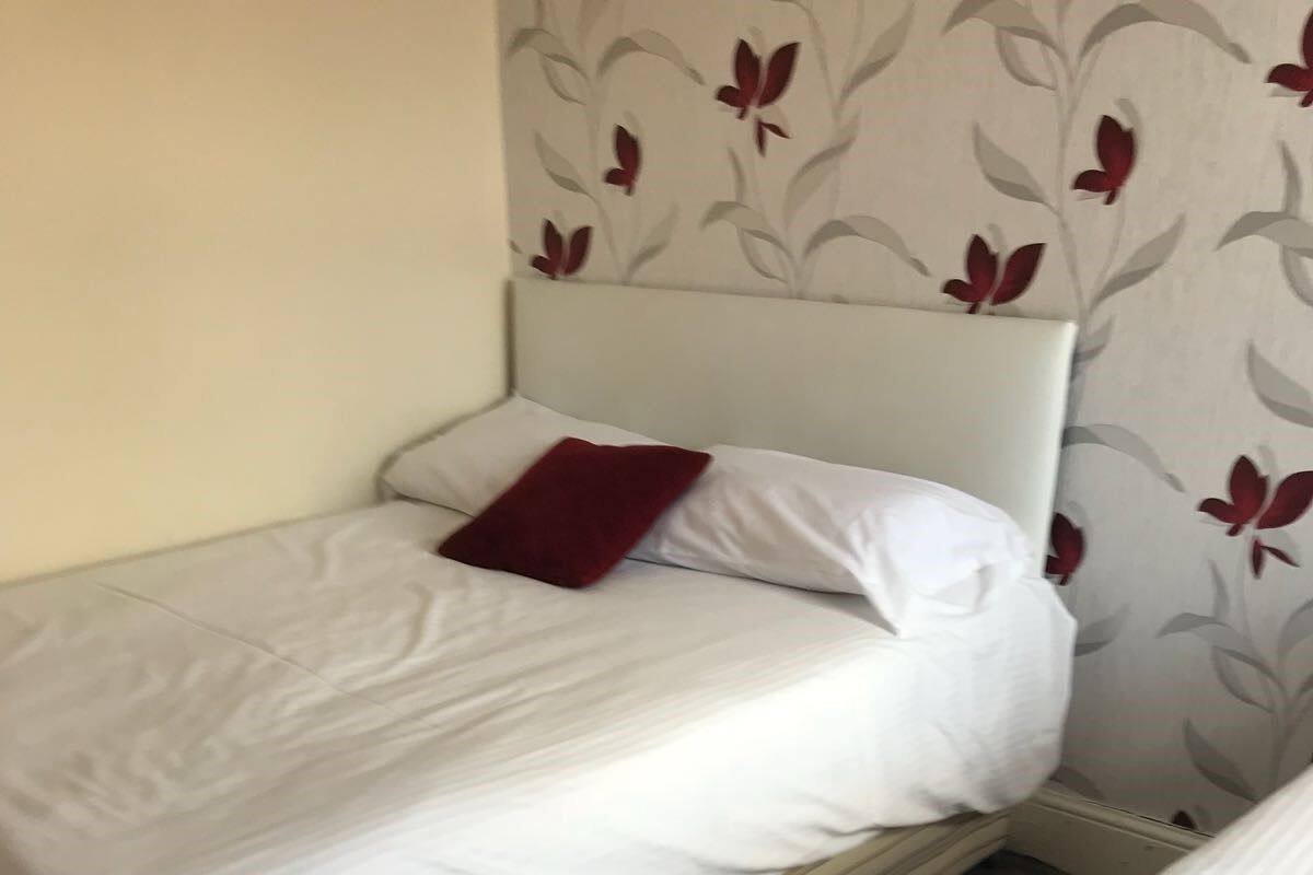 Una cama individual pequeña y bien hecha, con ropa de cama blanca y un cojín rojo, apoyada contra una pared con estampado de flores.