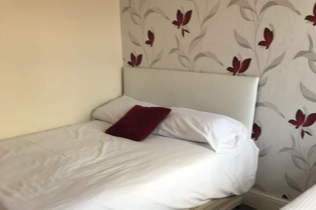 Uma cama de casal pequena e bem arrumada, com roupa de cama branca e um travesseiro vermelho, apoiada em uma parede com um papel de parede estampado com flores.