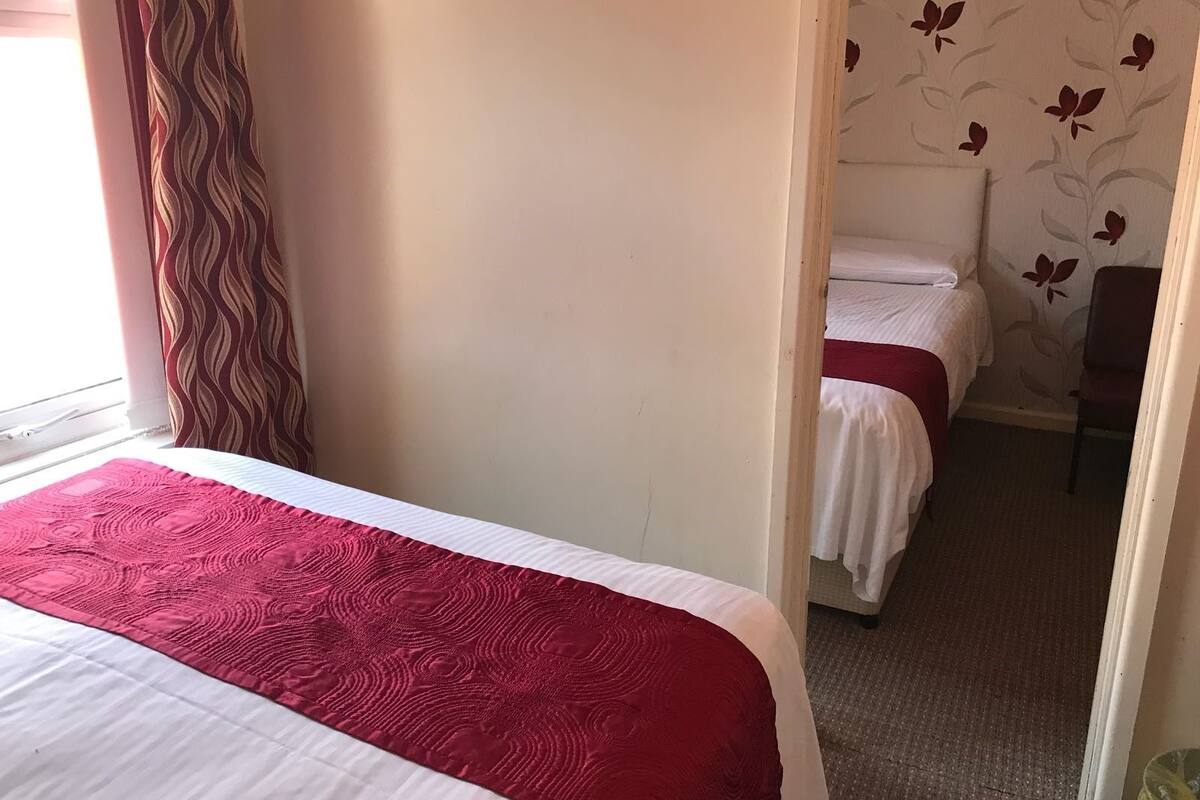 Quarto de hotel com duas camas, cada uma com colcha vermelha e branca, cortina estampada e papel de parede com motivos florais.