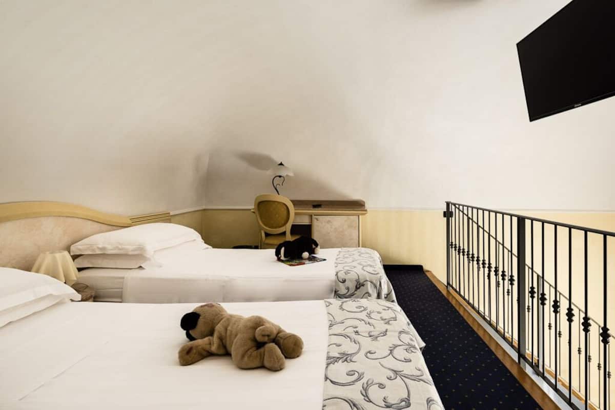 A hotel room with two beds, a desk, a chair, a black cat on one bed, and a teddy bear on the other.