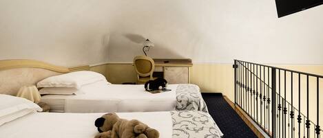 A hotel room with two beds, a desk, a chair, a black cat on one bed, and a teddy bear on the other.