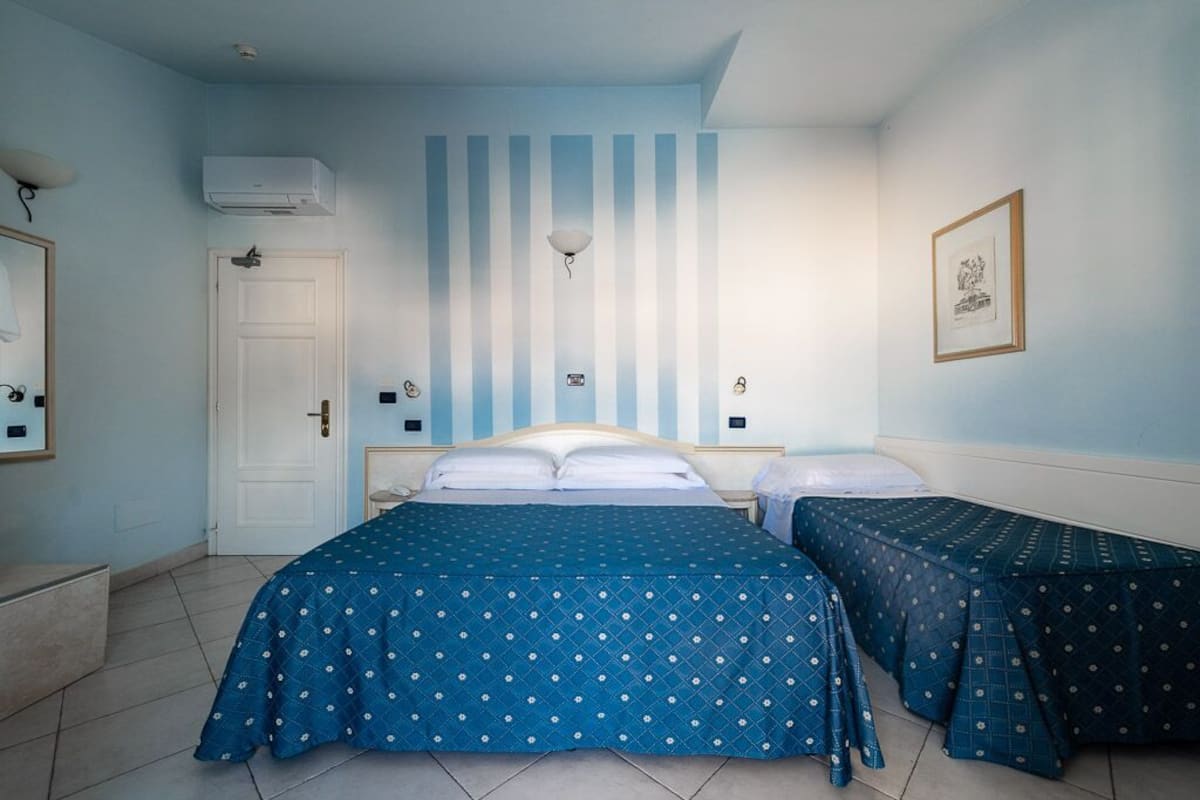 A room with a large bed, two smaller beds, a door, an air conditioning unit, a wall-mounted light, and a framed picture.
