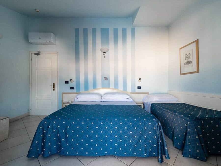 A room with a large bed, two smaller beds, a door, an air conditioning unit, a wall-mounted light, and a framed picture.