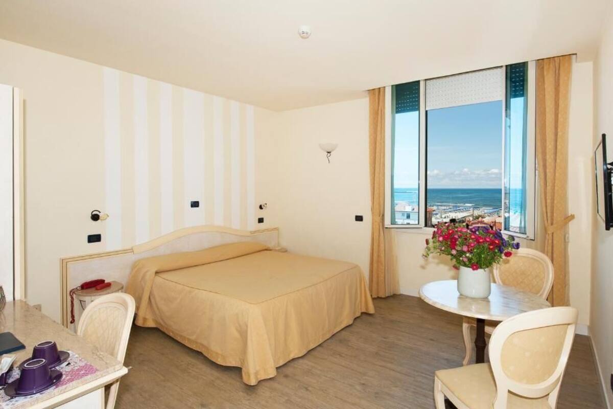 A hotel room with a bed, a small table with chairs, a desk with a telephone, and a window with a view of the sea.