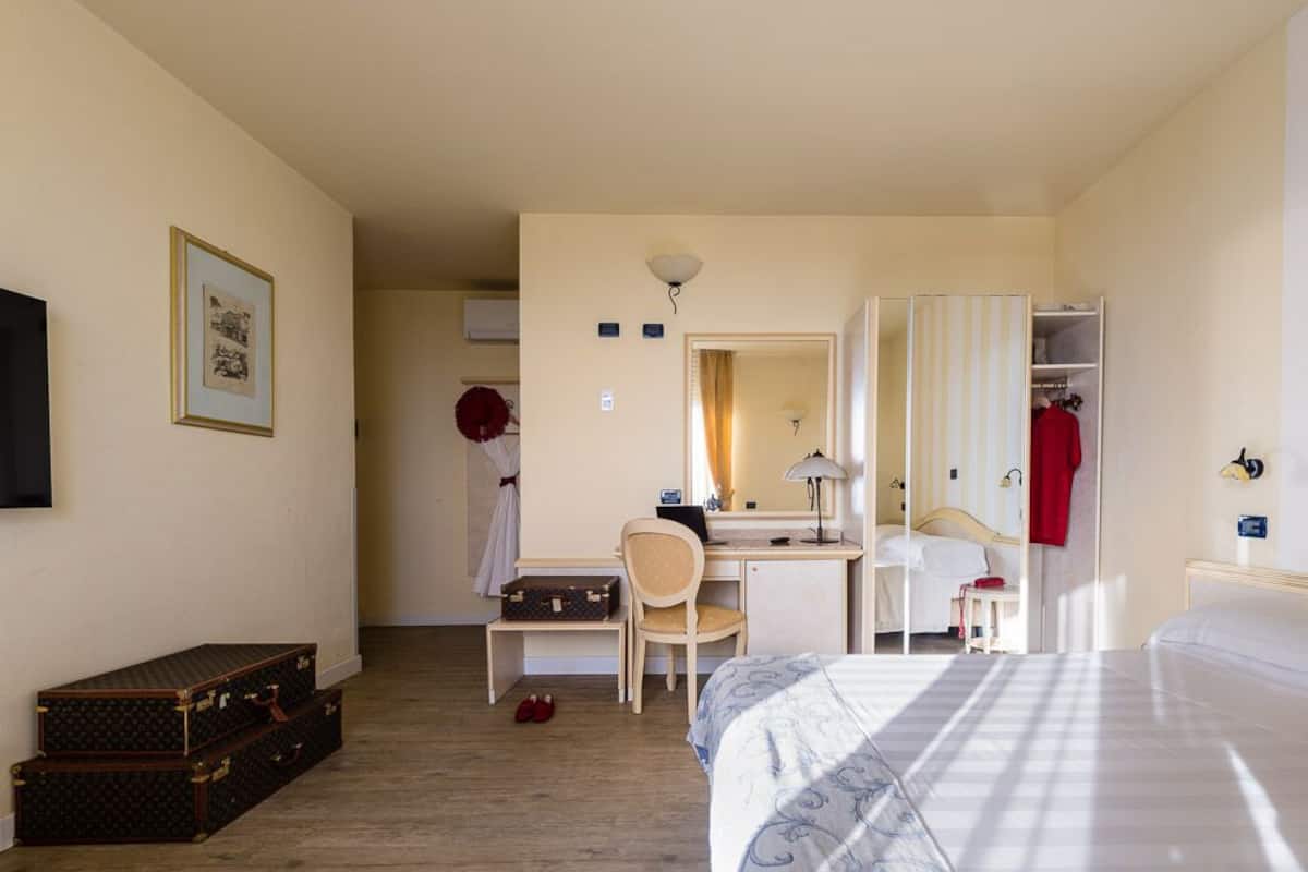 A hotel room with a bed, desk, chair, wardrobe, and a small table with a lamp.