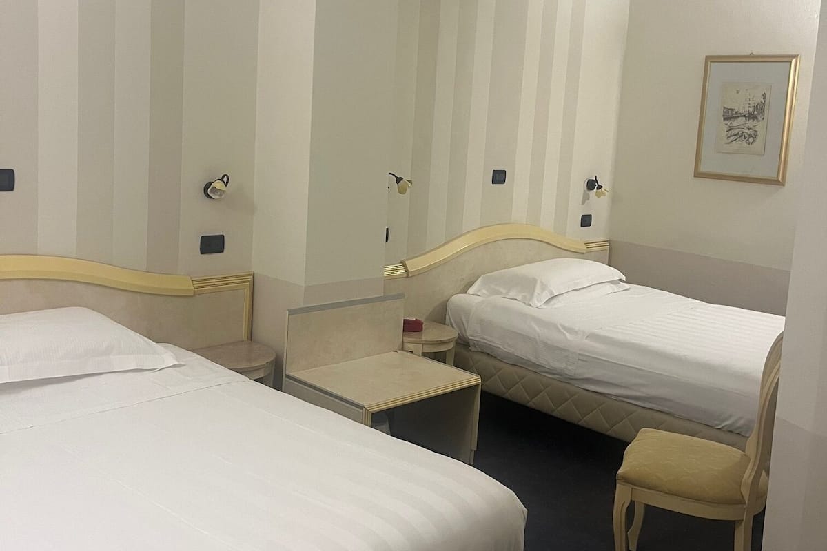 A hotel room with two single beds, a small bedside table, a chair, and a framed picture on the wall.