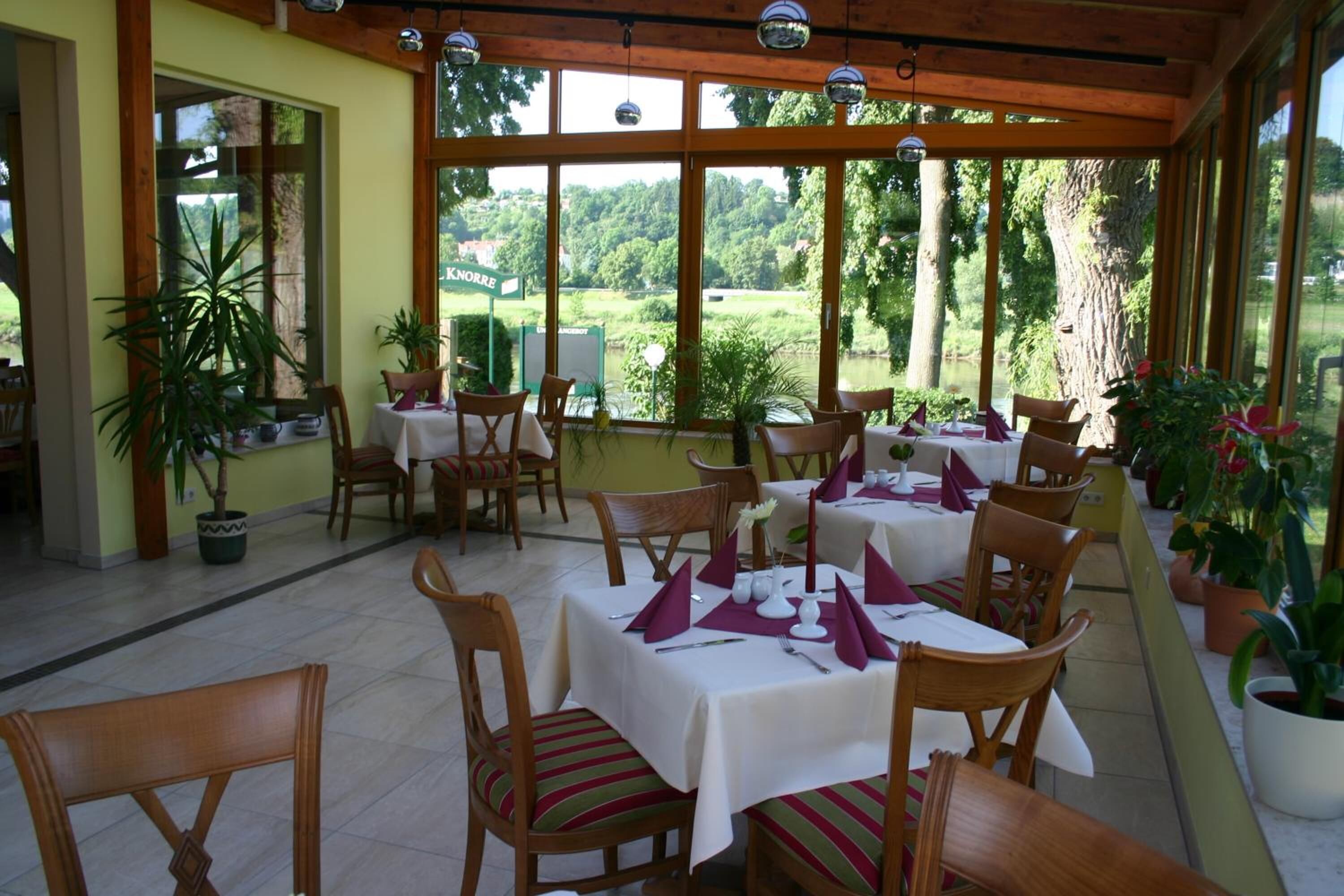 A restaurant with outdoor seating, wooden tables, and chairs, surrounded by large windows offering a view of greenery.