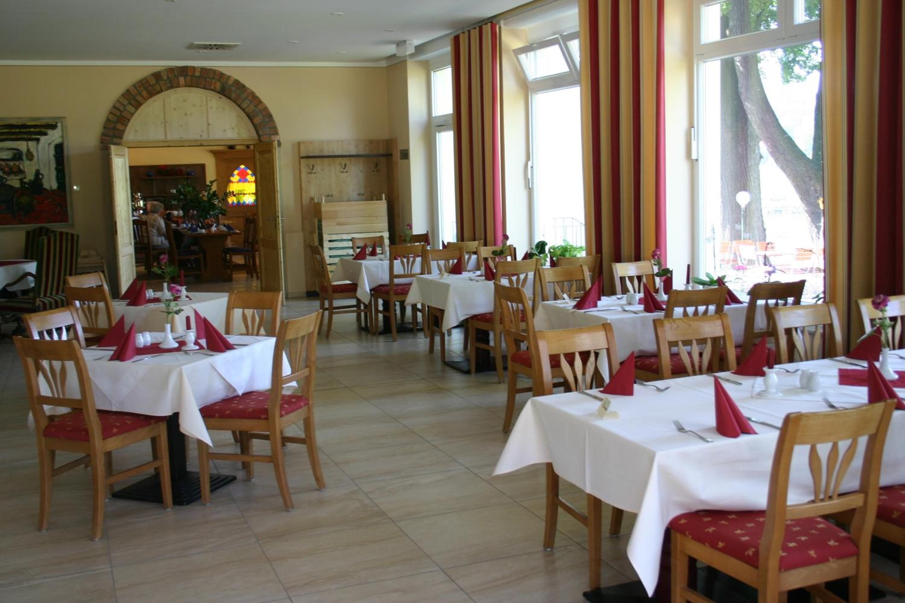 A dining area with multiple tables and chairs, a brick archway, and large windows with curtains.