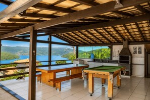 A pool table and a billiards table in a room with large windows overlooking a lake and hills.