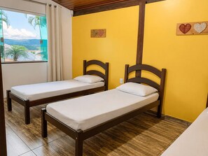 Duas camas de solteiro com estrutura de madeira em um quarto com paredes amarelas e uma janela com vista para palmeiras.