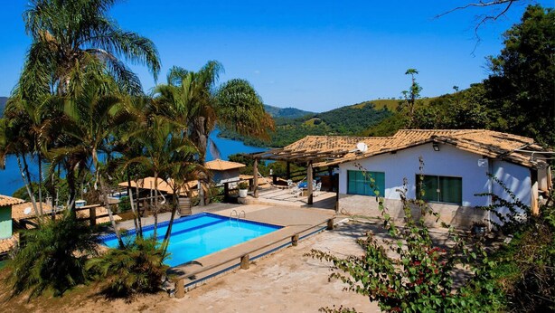 A pool area with a clear view of the sea, surrounded by palm trees and a small building with a tiled roof.