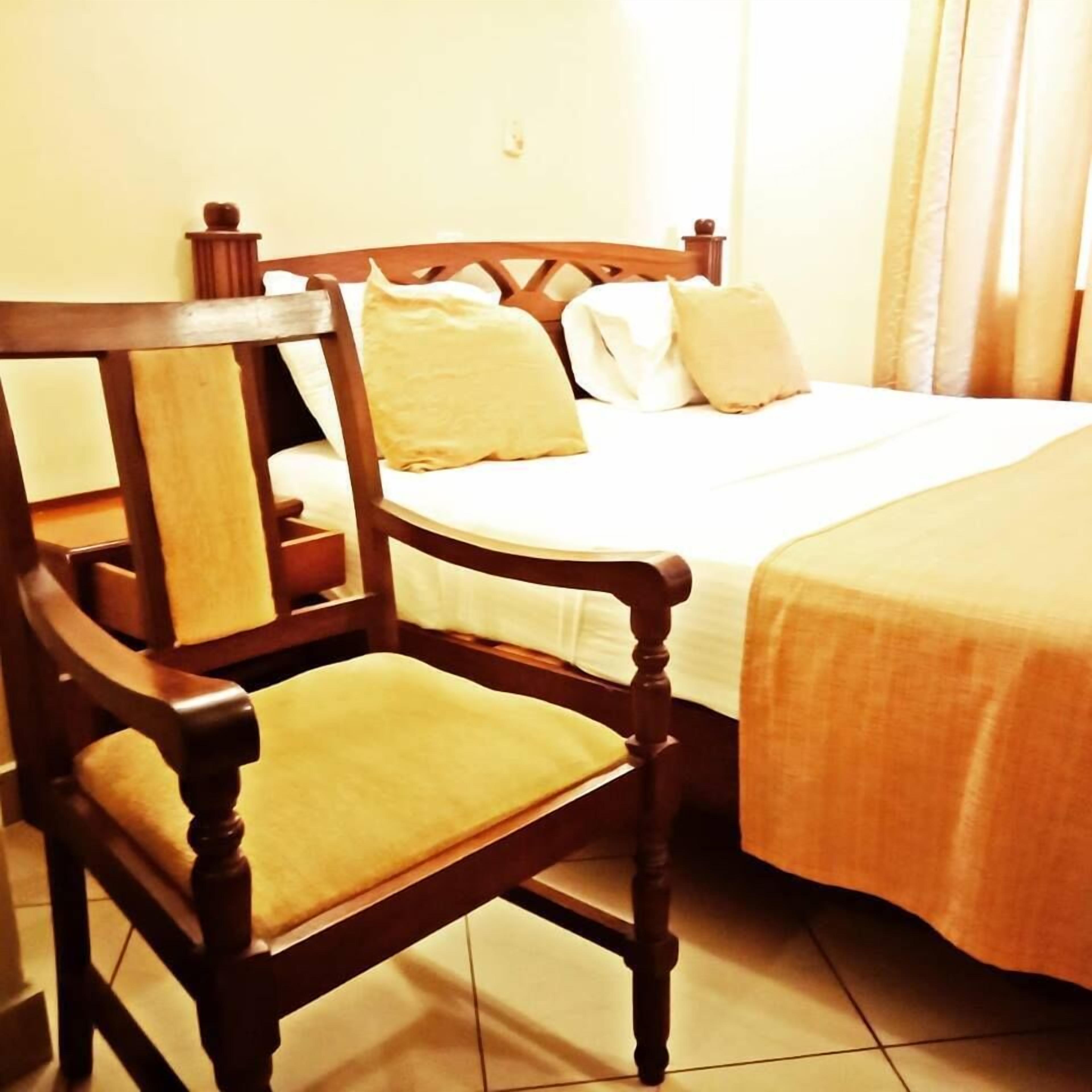 Una sedia in legno con un cuscino, un letto con materasso bianco e due cuscini, e una coperta beige.
