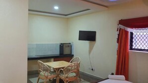 Una habitación pequeña con una cama, una mesa con sillas, un televisor montado en la pared y un ventanal con cortinas.