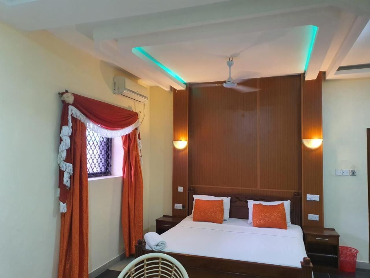 Une chambre d’hôtel comprenant un lit, une tête de lit en bois, deux tables de chevet, une fenêtre avec des rideaux, un ventilateur de plafond et des lampes fixées au mur.