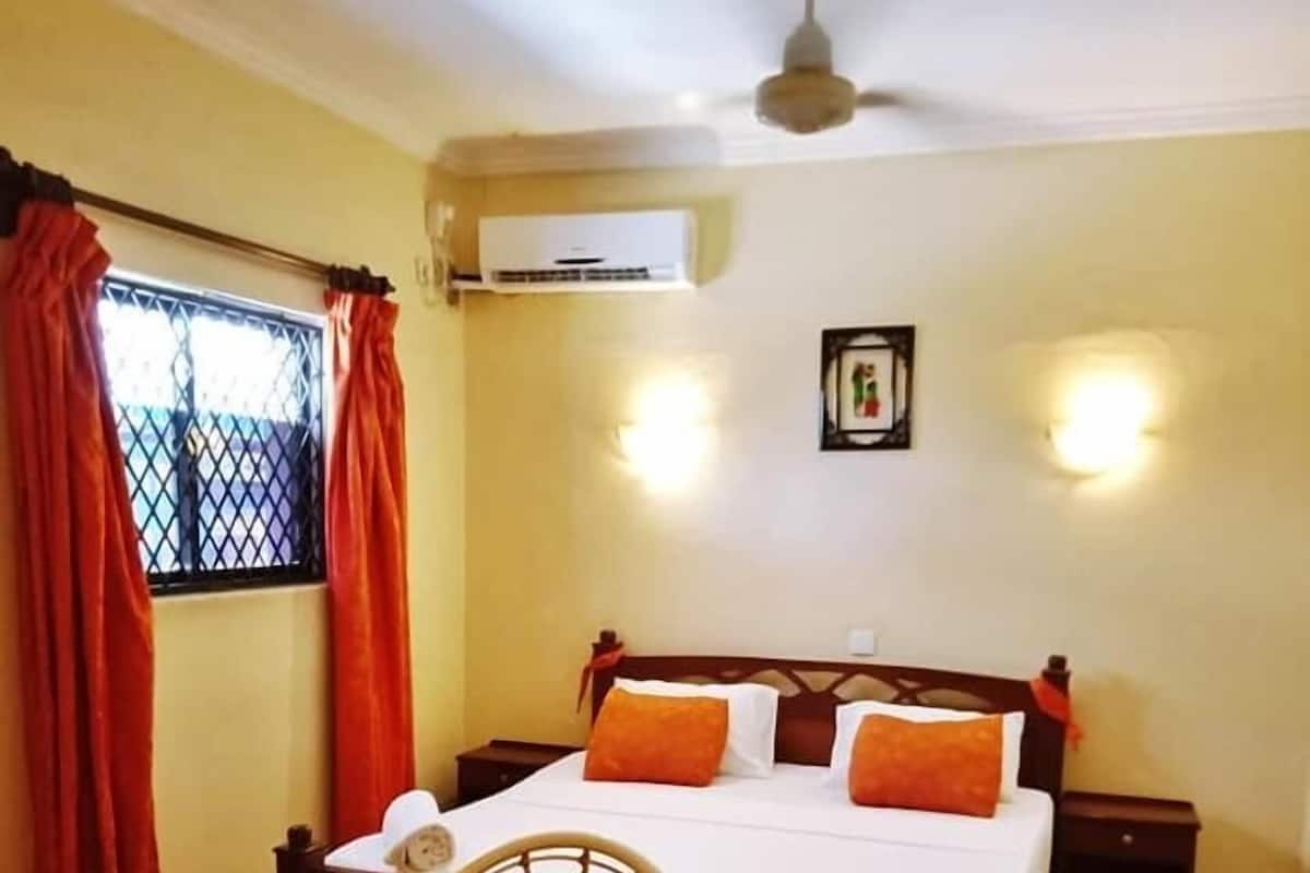 A room with a bed, a table, chairs, an air conditioning unit, and a wall-mounted light fixture.