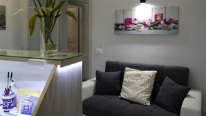 A room with a glass-top desk, a dark grey sofa with patterned cushions, and a wall-mounted light fixture. - Bed & Breakfast Conte Perollo (Sciacca)