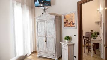 Una habitación con televisión montada en la pared, un armario blanco, un mueble pequeño, un cuadro enmarcado y una zona de comedor visible a través de una puerta abierta.