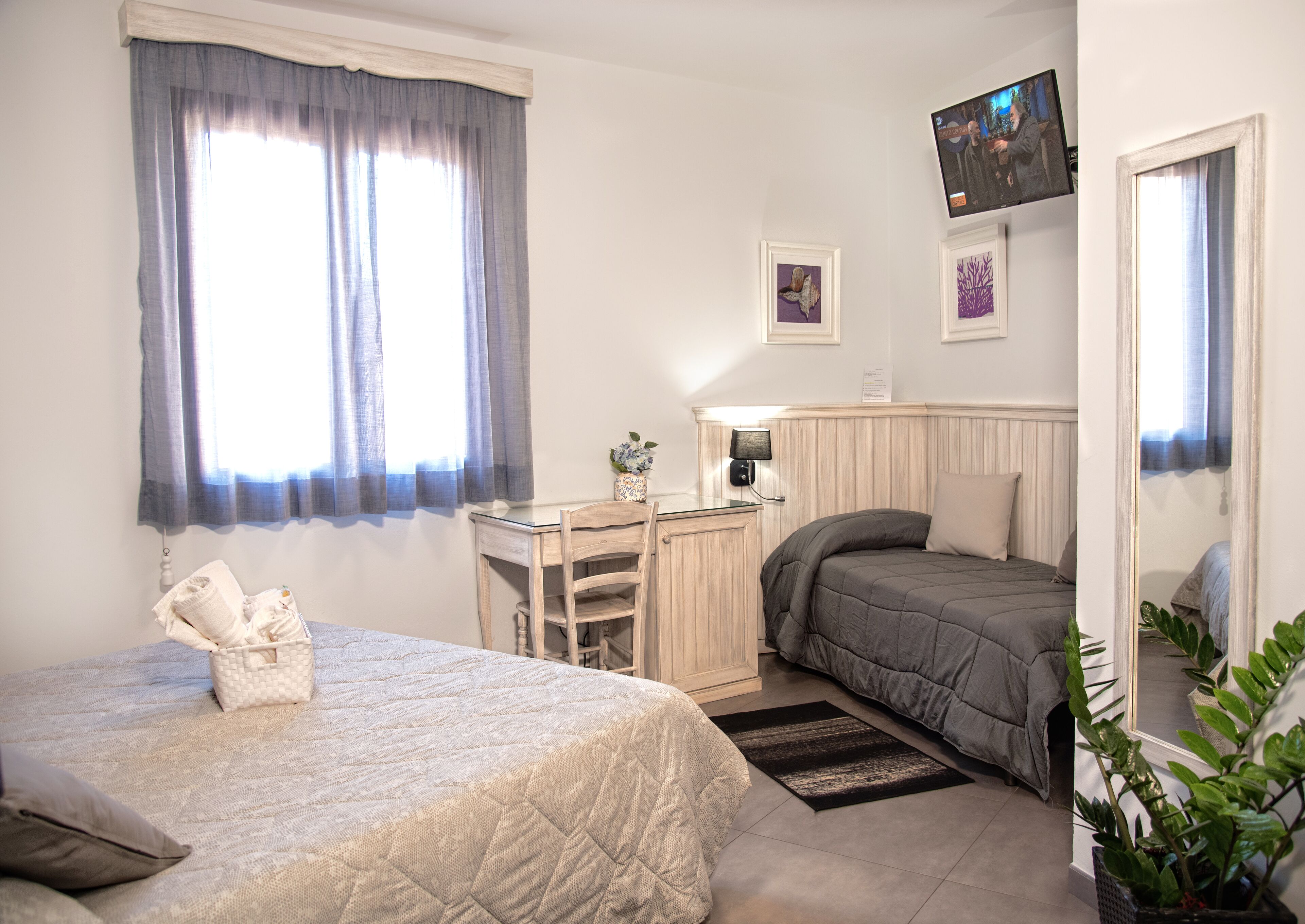 A bedroom with a bed, a desk, a chair, a bedside table, a lamp, a mirror, and a window with curtains.