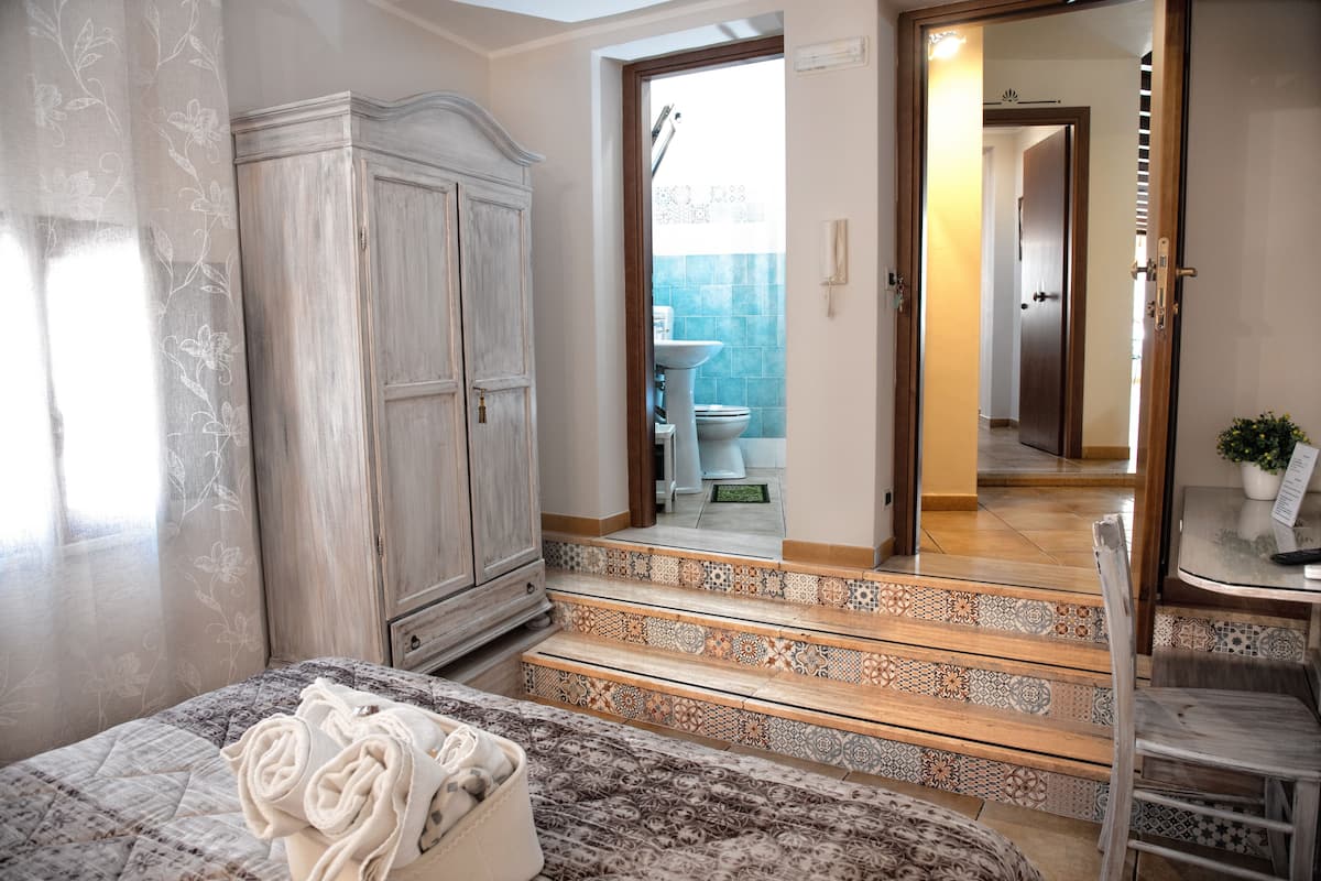 A bedroom with a bed, a wardrobe, a bathroom visible through an open door, and a small table with a plant.
