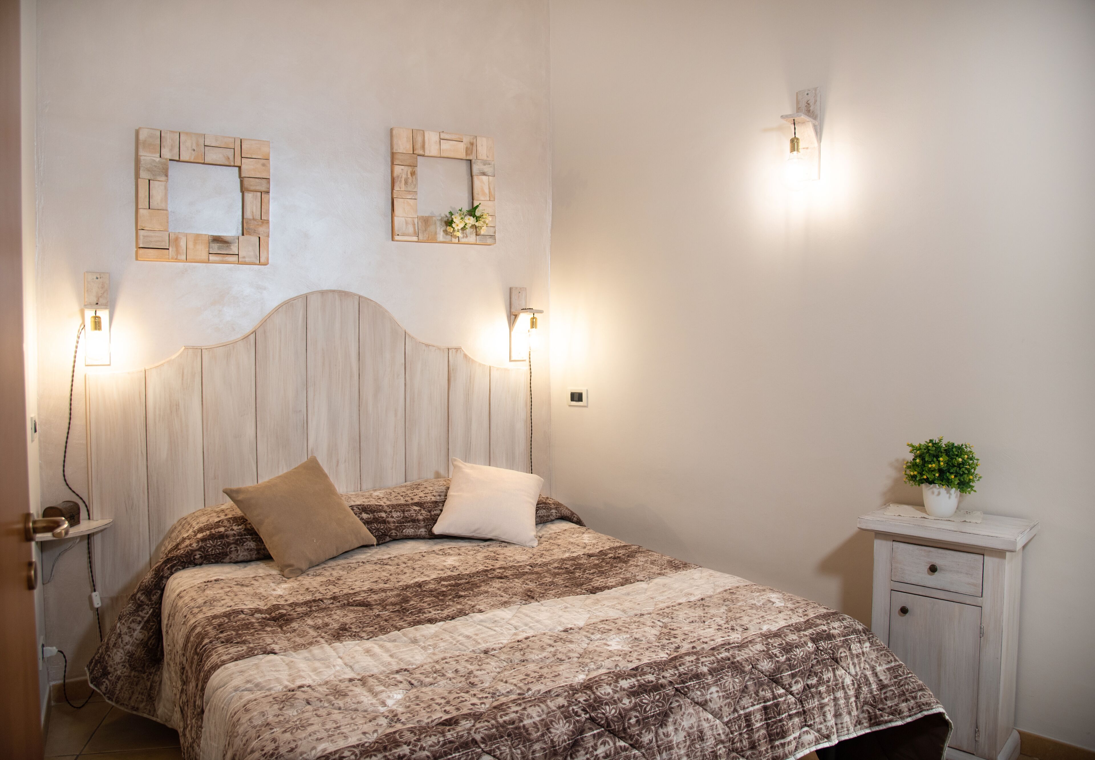 Un dormitorio con cabecera de madera, colcha estampada, dos luces fijadas a la pared, una mesita de noche con una planta y una mesita auxiliar con lámpara.