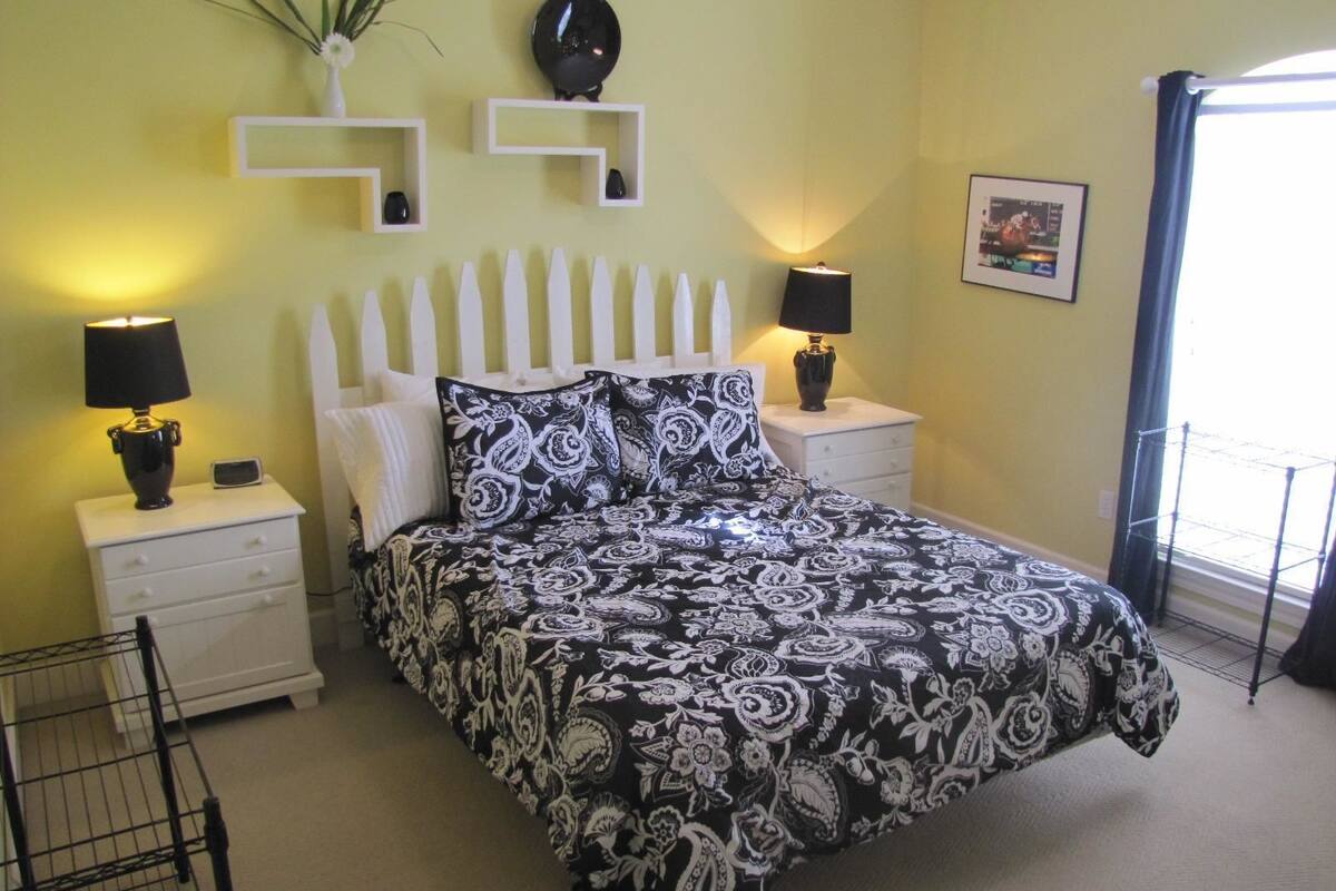 A bedroom with a bed, two bedside tables, two lamps, a wall clock, a framed picture, and a window with a curtain.