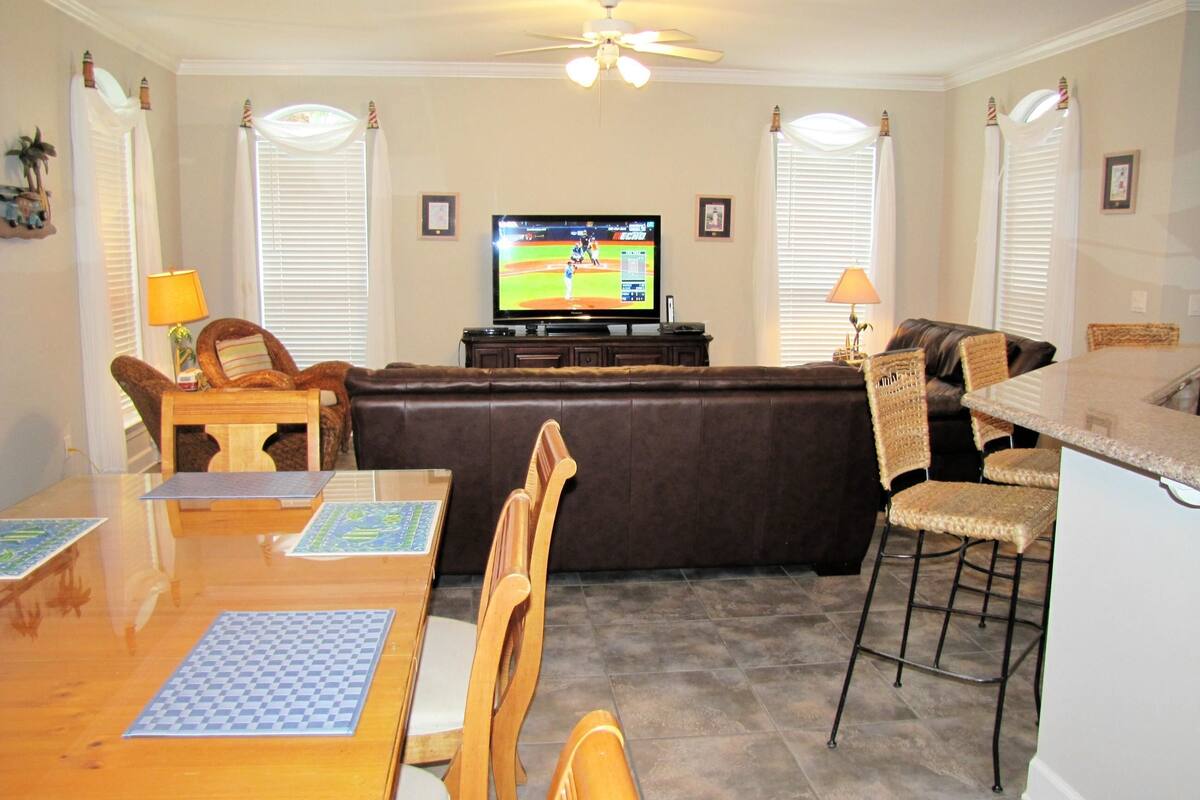 A living room with a television, a brown leather couch, a dining table with chairs, and a kitchen area with bar stools.