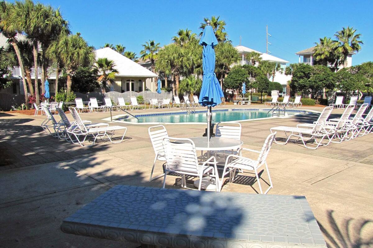 A poolside area with lounge chairs and umbrellas, surrounded by palm trees and buildings.