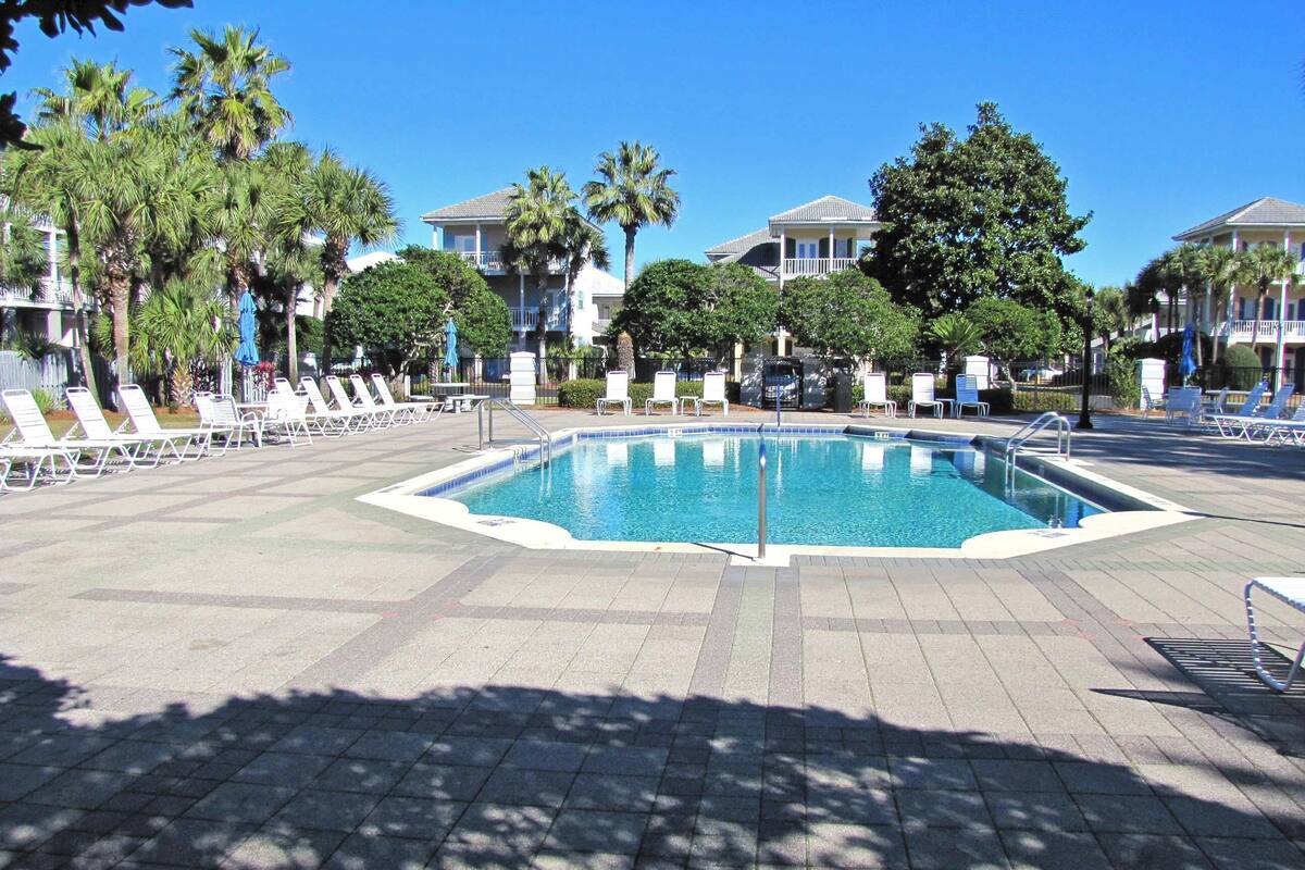 A swimming pool surrounded by lounge chairs and palm trees, with buildings in the background.