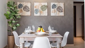 A dining area with a wooden table set for four, surrounded by white chairs. There are three framed artworks on the wall above the table, depicting stylized leaves. A large potted plant is visible to the left of the table. - Maison Privee - Exclusive Apt w/ Burj Khalifa Next Door (Dubai)
