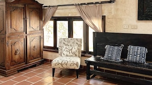 Une pièce comprenant un meuble en bois, un fauteuil à motifs, un canapé foncé avec des coussins à motifs, un luminaire suspendu au plafond et une fenêtre avec des rideaux.