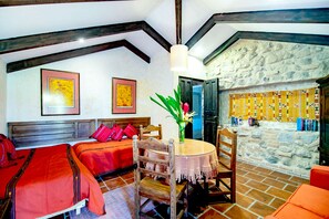 A room with a bed, a table with chairs, a vase with flowers, and a stone wall with a patterned cloth.
