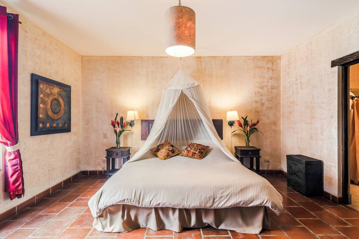 Une chambre à coucher avec un lit sous un dais, deux tables de chevet avec des lampes, un rideau rouge et une œuvre d’art accrochée au mur.