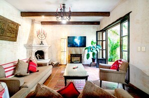 A living room with a fireplace, sofa, armchair, television, and a large window with plants.