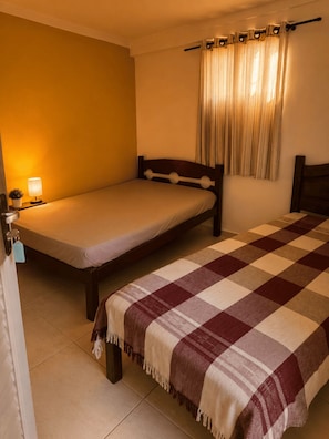 Una cama individual con cabecera de madera, una lámpara de noche, una manta a cuadros y una ventana con cortinas.