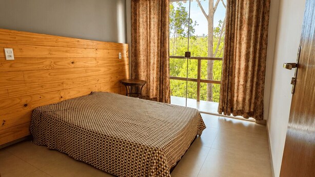 A bedroom with a wooden headboard, a bed with a patterned bedspread, a small round table, and a window with curtains. -  Paz e Tranquilidade - Casa para 20 pessoas com 7 quarto (Capitólio)