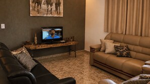 A living room with a leather sofa, a television on a wooden stand, a painting of horses, and a curtain. -  Paz e Tranquilidade - Casa para 20 pessoas com 7 quarto (Capitólio)