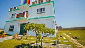 Um edifício de dois andares com uma porta verde, janelas e uma varanda com guarda-corpo. Há áreas de estar ao ar livre e uma pequena árvore no jardim da frente.