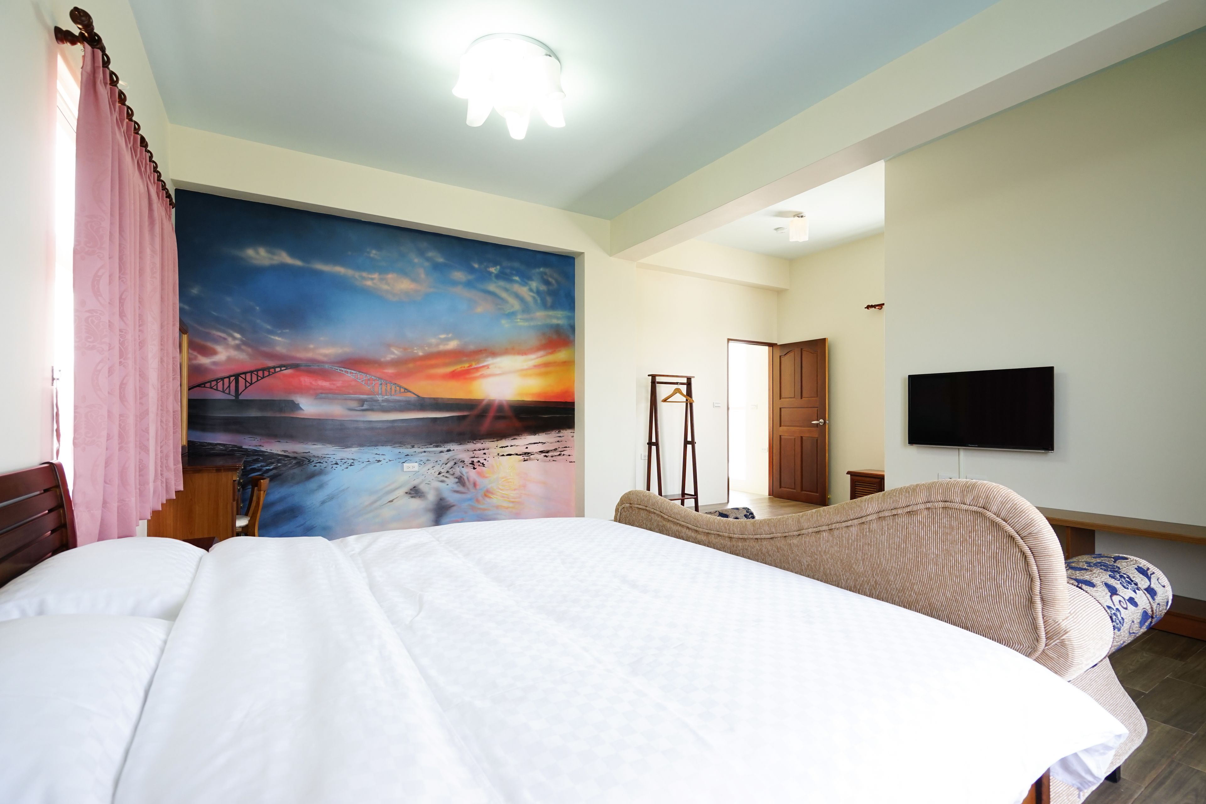 A bedroom with a bed, a large painting of a sunset, a TV, and a wooden chair.