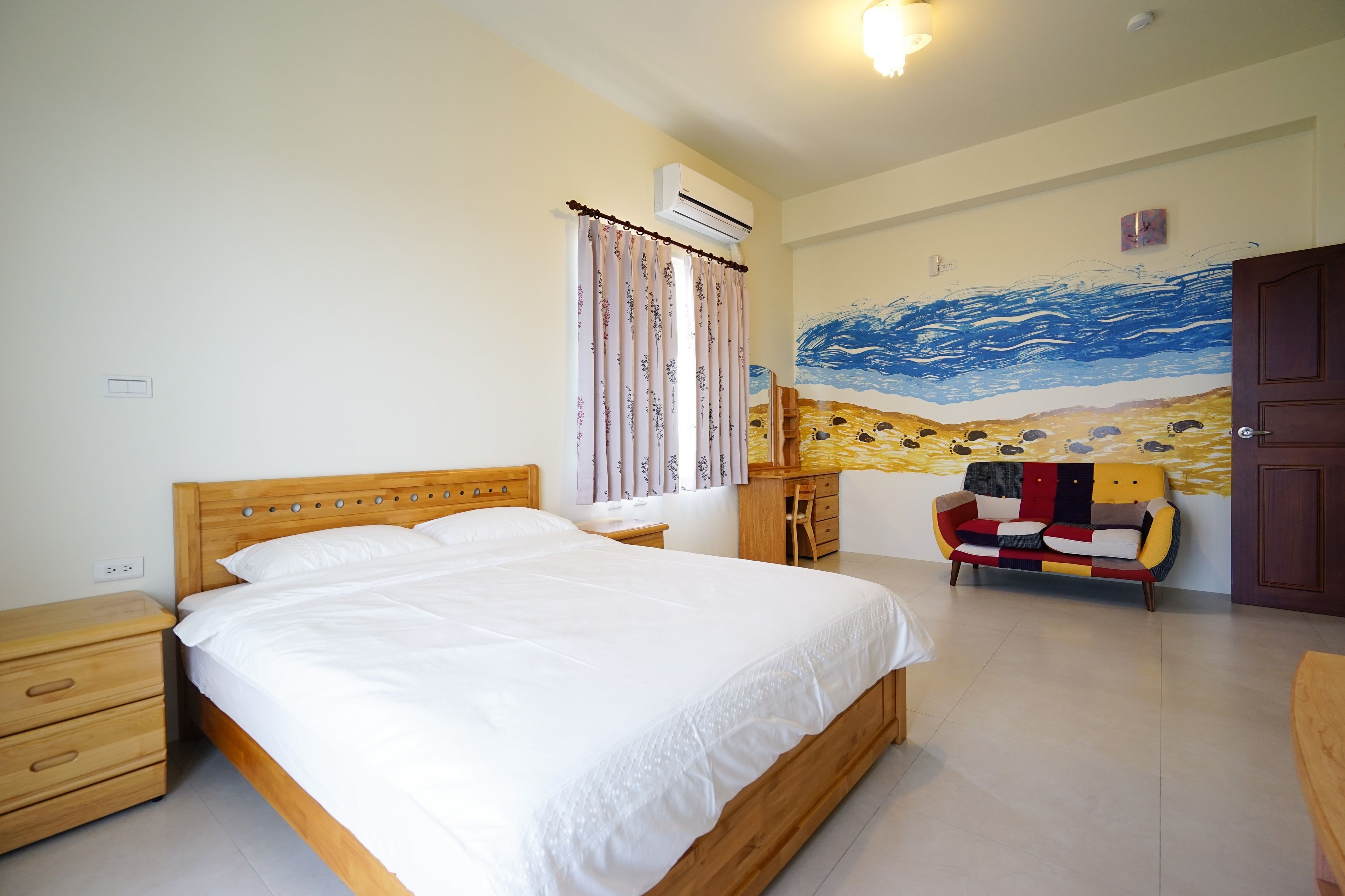 A bedroom with a wooden bed, a nightstand, a dresser, a colorful sofa, and a wall mural.
