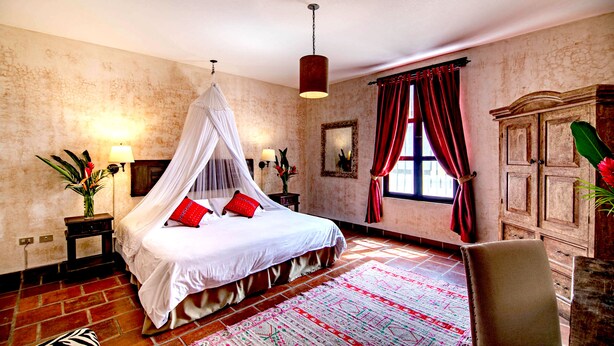 Une chambre à coucher avec un lit à baldaquin, un tapis aux motifs rouges et blancs, une armoire en bois et une grande fenêtre ornée de rideaux rouges.