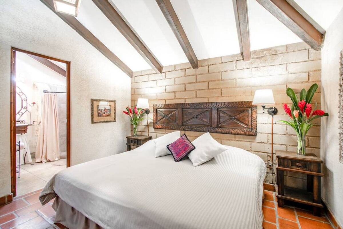Een slaapkamer met een groot bed, houten balken, bakstenen muren en twee nachtkastjes met lampen en bloemen.