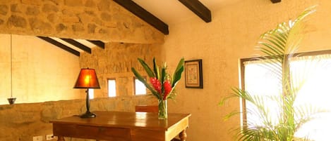 Una stanza con parete in pietra, travi in legno, pavimento in piastrelle, un tavolo in legno con un vaso di fiori e una pianta in vaso.