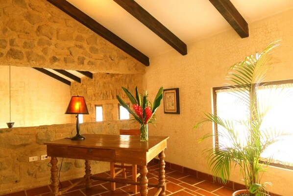 Une pièce avec un mur en pierre, des poutres apparentes, un sol carrelé, une table en bois ornée d’un vase de fleurs et une plante en pot.