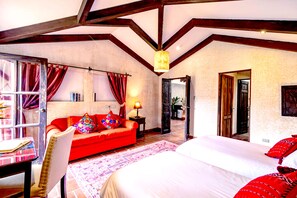 Une pièce spacieuse avec un grand lit, un canapé rouge, un fauteuil et une table avec des livres. Il y a une porte qui donne sur une autre pièce et une fenêtre avec des rideaux.